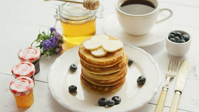 From above view of pancakes served in white plate decorated with slices of banana and blueberries with cup of coffee and honey placed near on white background.